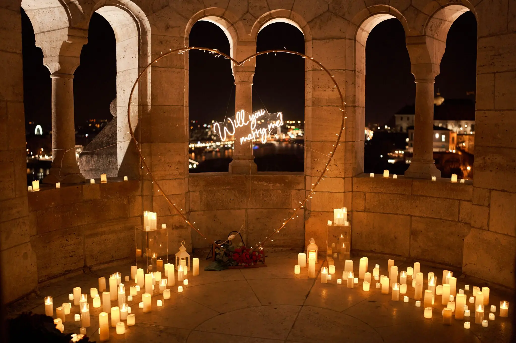 Marriage proposal setup in Romania — candlelit heart arch with "Will you marry me?" sign, stone arches and city lights in the background